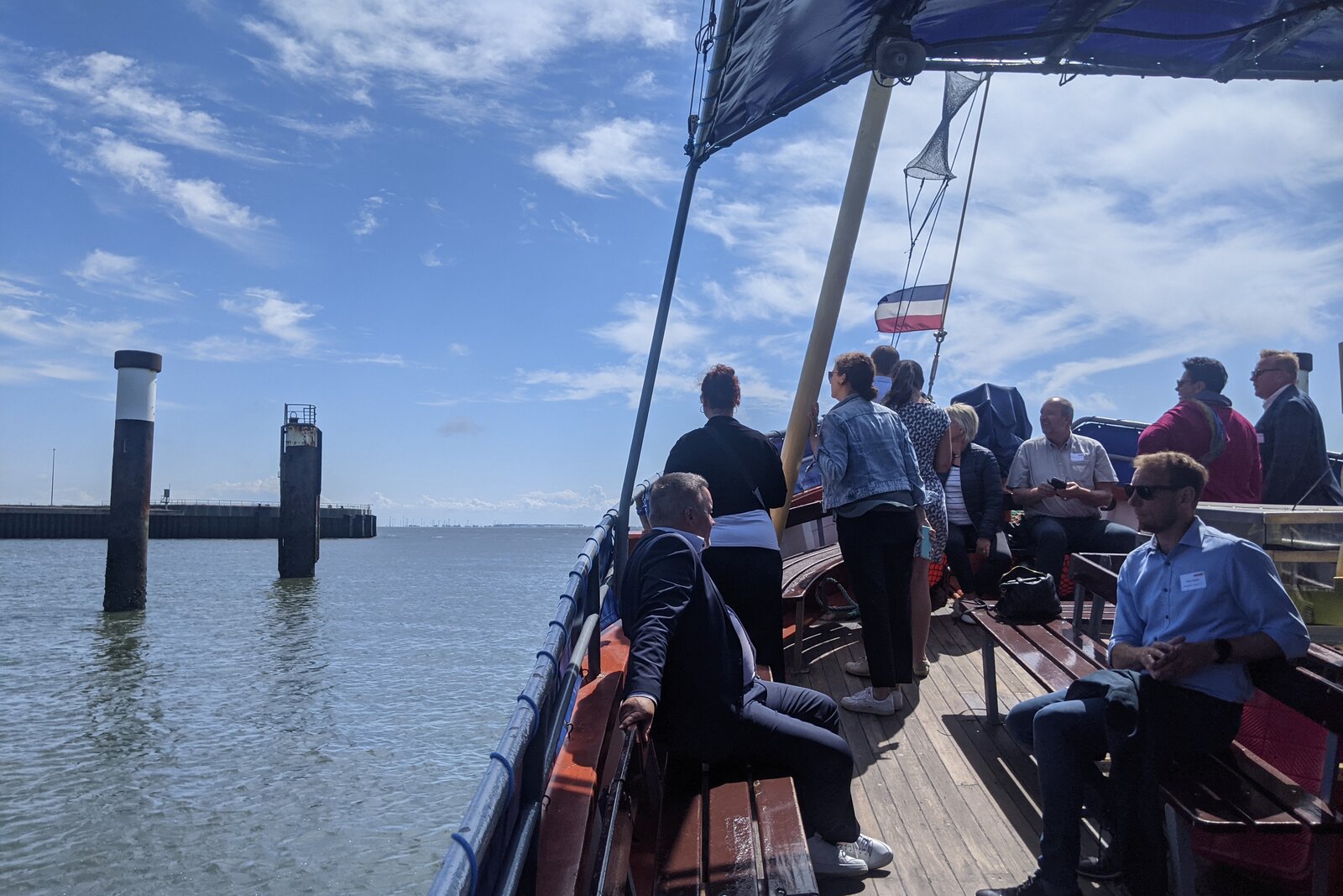 Eine Gruppe von Menschen befindet sich auf einem Bootsausflug bei sonnigem Wetter. Die Passagiere stehen oder sitzen auf dem hölzernen Deck, einige unterhalten sich, während andere die Aussicht auf das Wasser genießen. Im Hintergrund sind Hafenstrukturen mit hohen Pfeilern sowie eine weite Wasserfläche unter einem blauen Himmel mit weißen Wolken zu sehen. Eine Fahne weht im Wind, und das Boot hat ein teilweise überdachtes Deck. Die Atmosphäre wirkt entspannt und gesellig.