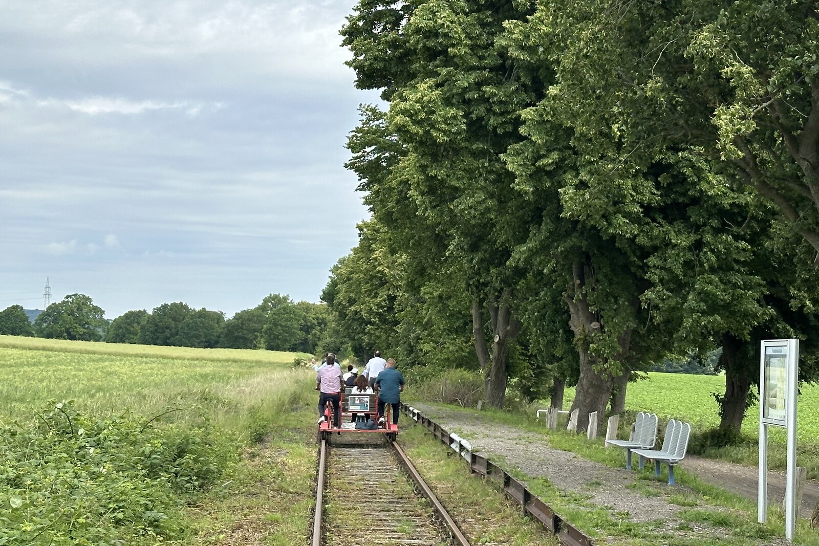 Eine Gruppe von Menschen f&auml;hrt mit einer Fahrraddraisine auf einem stillgelegten Bahngleis durch eine l&auml;ndliche Landschaft. Rechts verl&auml;uft eine Baumreihe mit einer kleinen Haltestelle, links liegen Felder unter einem bew&ouml;lkten Himmel.
