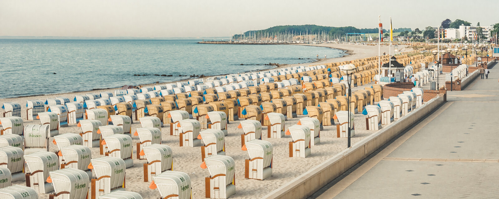 Das Bild zeigt eine breite Strandpromenade an einem ruhigen Meeresufer bei klarem Himmel. Auf dem weißen Sandstrand stehen in gleichmäßigen Reihen zahlreiche Strandkörbe in Weiß und Braun, viele davon mit Aufschriften oder kleinen Markierungen. Die Körbe sind größtenteils leer. Rechts verläuft eine gepflegte Promenade mit Laternen, Fahnenmasten und kleinen Häuschen. Im Hintergrund erstreckt sich eine Kurve des Strandes bis zu einem Yachthafen mit vielen Masten. Dahinter ist eine grüne, bewaldete Landzunge zu sehen. Einige Spaziergänger sind auf der Promenade unterwegs. 