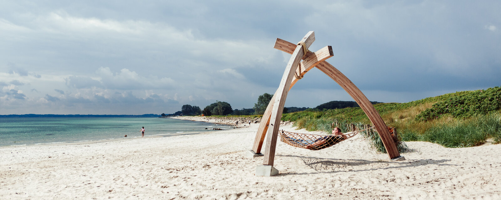 Person liegt in einer Hängematte unter einer modernen Holzstruktur am weißen Sandstrand, mit Blick aufs Meer und unter leicht bewölktem Himmel.