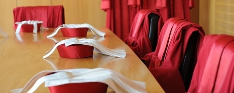 Ein Konferenzraum mit einem langen Holztisch, auf dem rote Richterhüte mit weißen Bändern liegen. Im Hintergrund hängen an einer Garderobe rote Roben mit weißen Kragen. Die Szene deutet auf eine juristische Institution hin, möglicherweise ein Gericht oder eine Verfassungsinstanz. Die Anordnung der Roben und Hüte vermittelt einen förmlichen und feierlichen Eindruck.