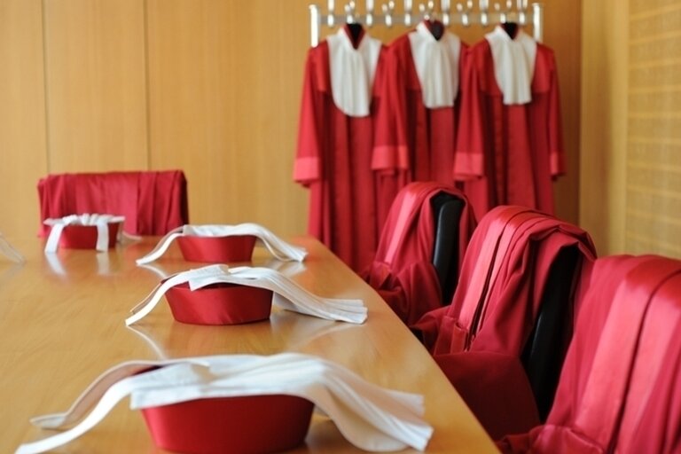 Ein Konferenzraum mit einem langen Holztisch, auf dem rote Richterhüte mit weißen Bändern liegen. Im Hintergrund hängen an einer Garderobe rote Roben mit weißen Kragen. Die Szene deutet auf eine juristische Institution hin, möglicherweise ein Gericht oder eine Verfassungsinstanz. Die Anordnung der Roben und Hüte vermittelt einen förmlichen und feierlichen Eindruck