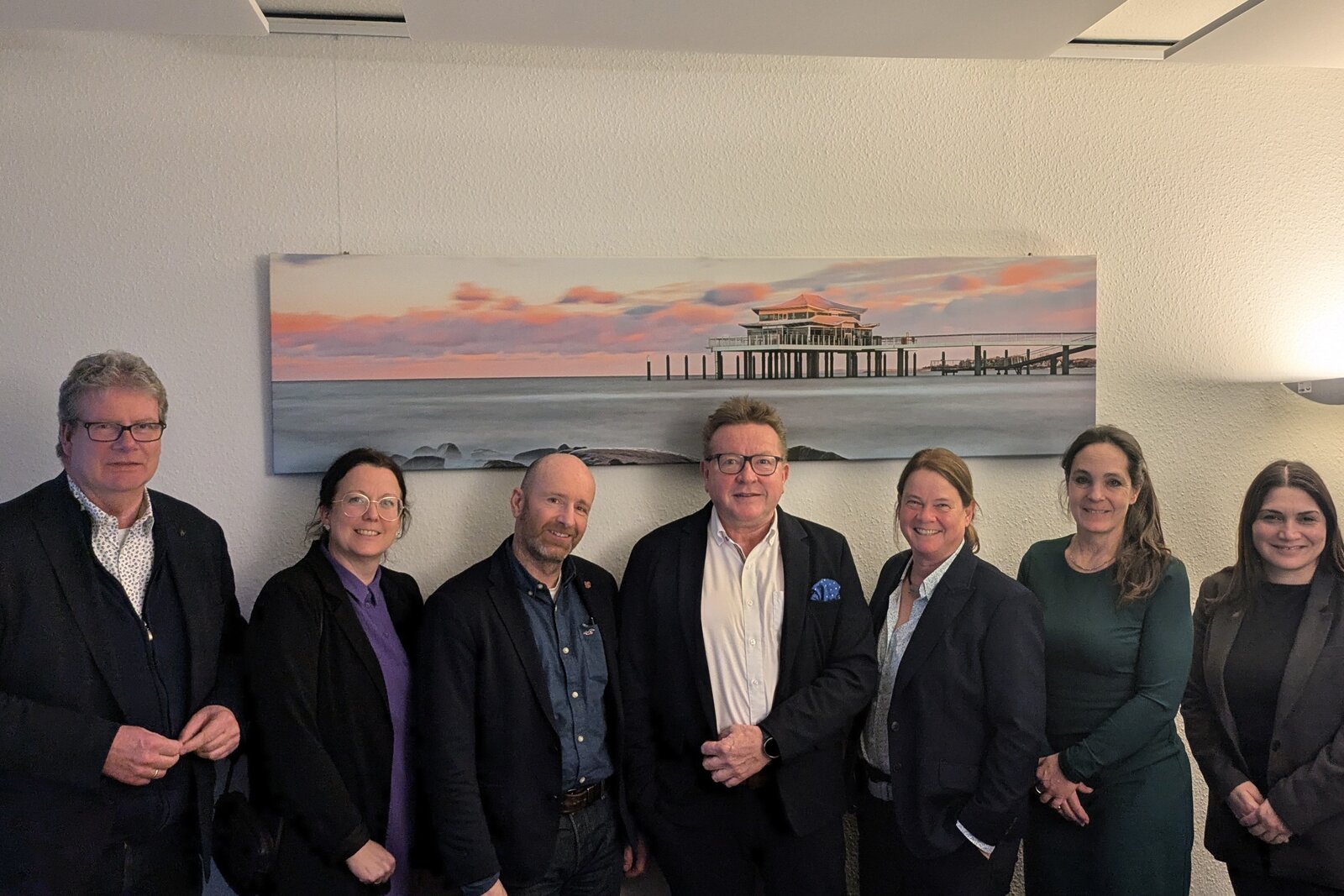 Das Bild zeigt eine Gruppe von sieben Personen, die in einem Büro oder Konferenzraum zusammenstehen. Alle Personen tragen geschäftliche Kleidung, darunter Blazer und Hemden. Die Gruppe besteht aus Männern und Frauen, die freundlich in die Kamera lächeln. Im Hintergrund hängt ein großes Bild an der Wand, das eine Seebrücke mit einem Gebäude darauf zeigt.
