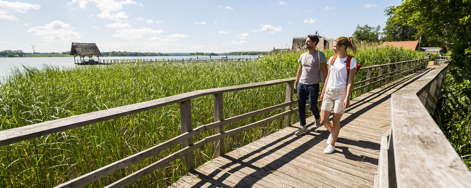 Das Bild zeigt einen sonnigen Sommertag an einem See mit einem hölzernen Stegweg, der durch ein Schilfgebiet führt. Zwei Menschen, ein Mann und eine Frau, spazieren entspannt auf dem Steg und unterhalten sich. Sie tragen sommerliche Kleidung und Rucksäcke. Links im Hintergrund erstreckt sich der See mit einem weiteren langen Holzsteg, der zu kleinen, mit Schilfdächern bedeckten Hütten führt. Der Himmel ist blau mit wenigen weißen Wolken, rechts säumen Bäume den Weg. Die Szene vermittelt Ruhe und Naturverbundenheit.