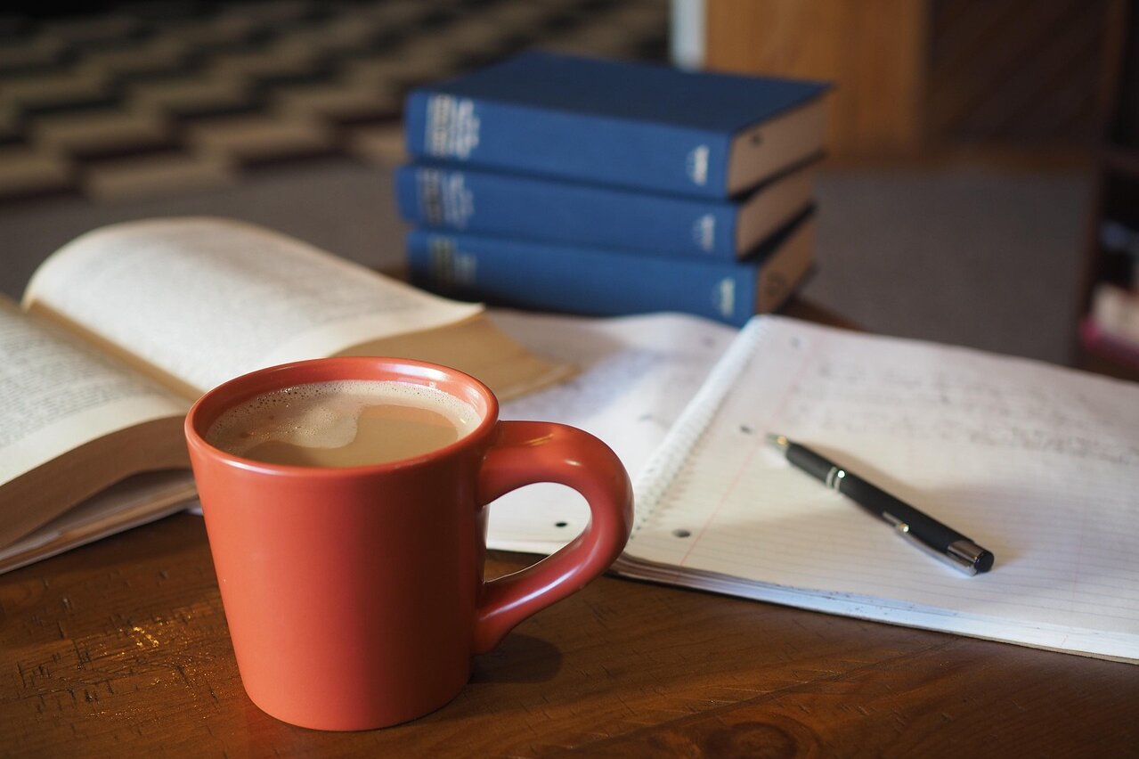 Auf dem Bild ist eine Kaffeetasse im Vordergrund zu sehen, die auf einem Tisch steht. Im Hintergrund ein aufgeschlagener Block und drei blaue Bücher.