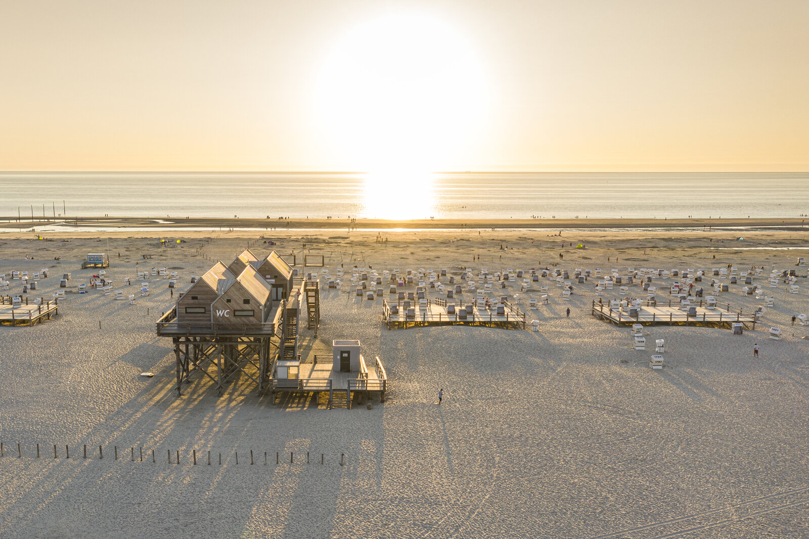 Ein malerischer Sonnenuntergang am Nordseestrand von St. Peter-Ording, mit den charakteristischen Pfahlbauten, zahlreichen Strandkörben und sanftem Abendlicht, das die Weite des Wattenmeers in goldene Farben taucht