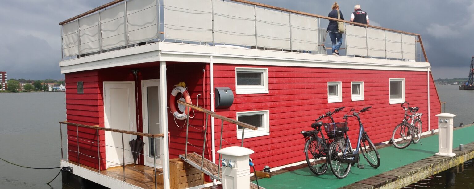 Ein modernes, rot gestrichenes Hausboot liegt an einem Holzsteg mit gr&uuml;ner Teppichauflage vor Anker. Das Hausboot hat eine wei&szlig;e T&uuml;r und mehrere rechteckige Fenster. Eine kleine Holztreppe mit einem Rettungsring f&uuml;hrt zum Eingang. Auf dem Dach befindet sich eine umz&auml;unte Terrasse, auf der zwei Personen stehen. Am Steg sind mehrere Fahrr&auml;der abgestellt, und das Wasser sowie ein bew&ouml;lkter Himmel sind im Hintergrund zu sehen.