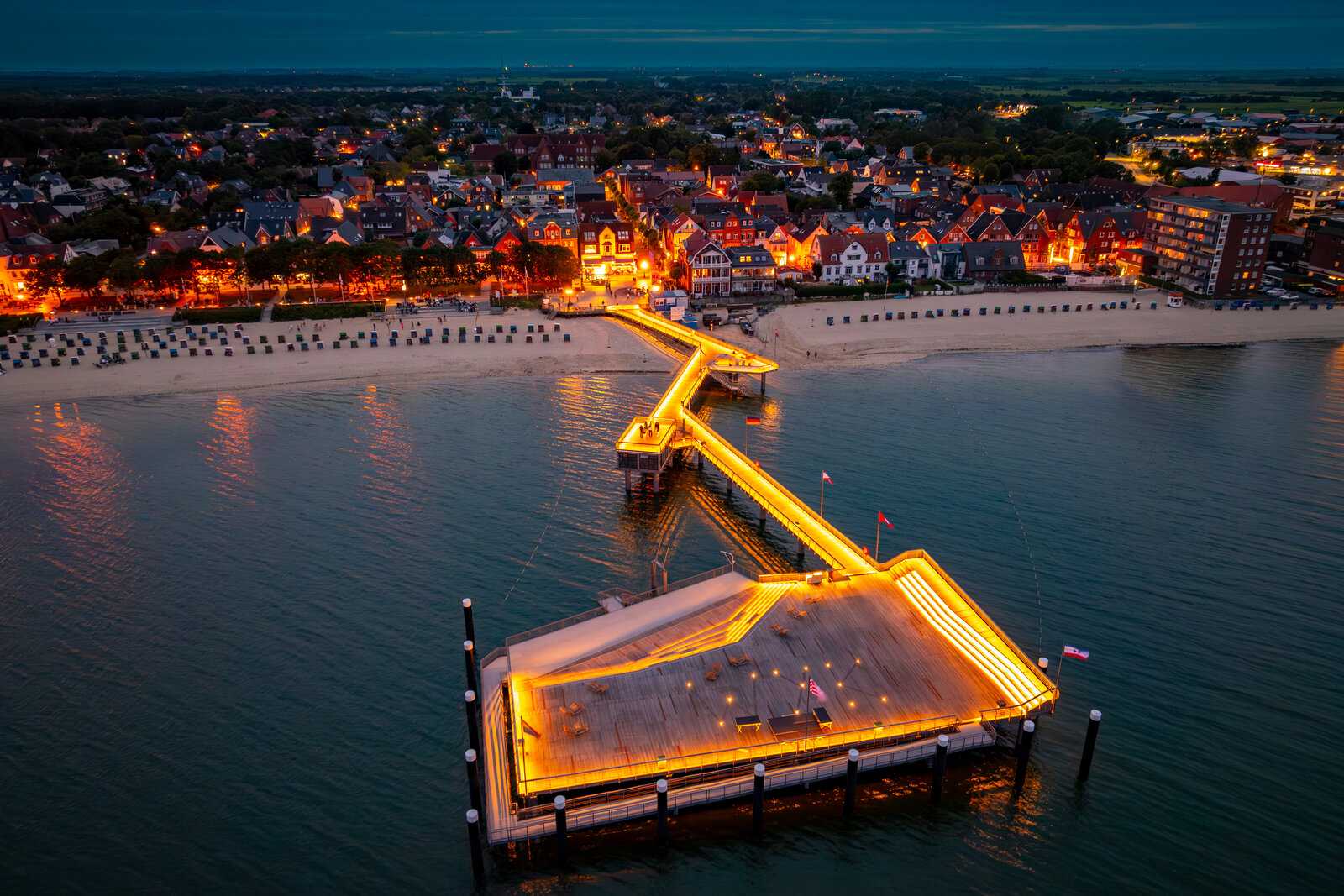 Die abendlich beleuchtete Seebrücke erstreckt sich strahlend ins Meer und verbindet die gemütlich erleuchtete Küstenstadt mit dem ruhigen Wasser. Strandkörbe säumen den Sandstrand, während die Lichter der Stadt eine stimmungsvolle Kulisse schaffen.
