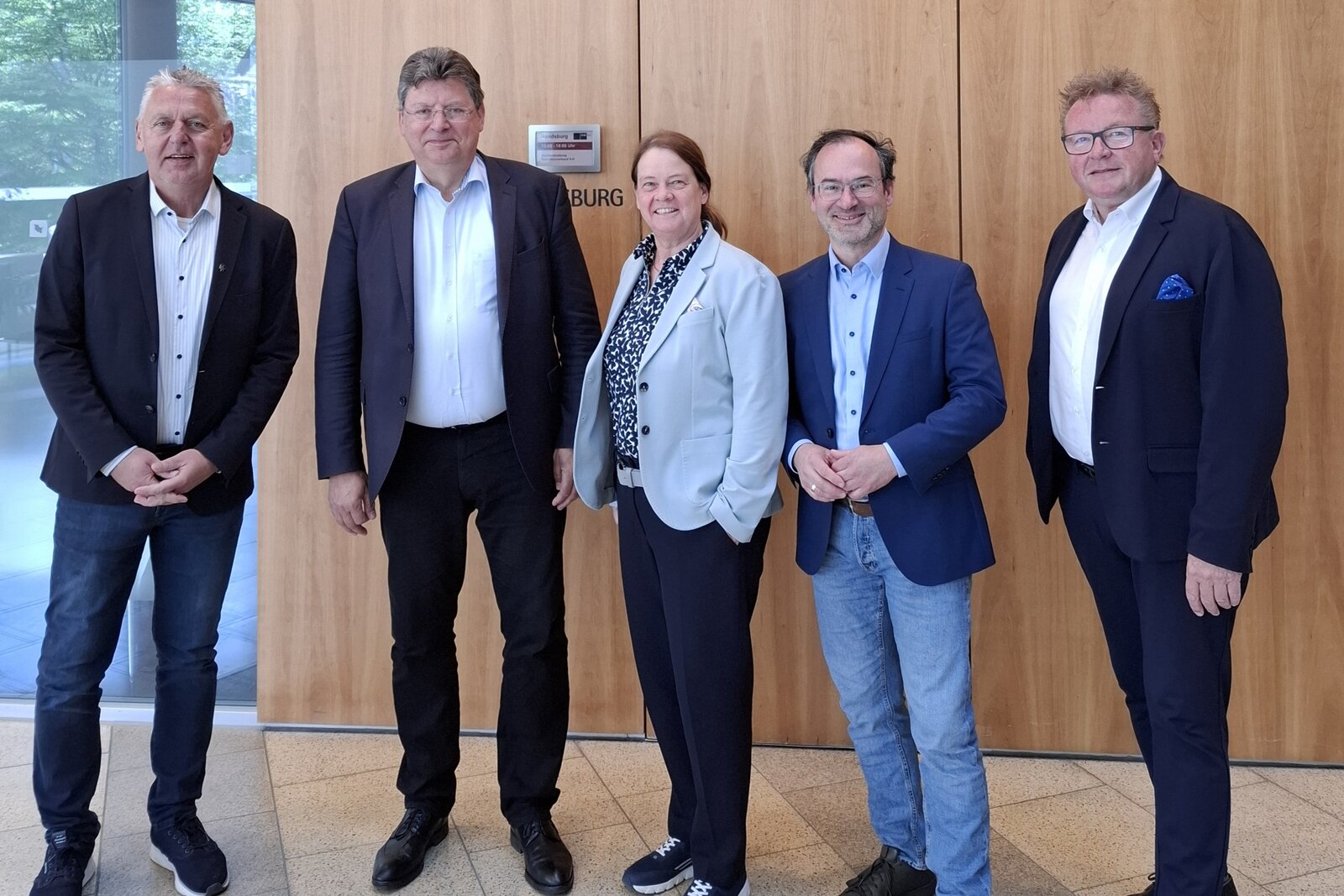 Das Bild zeigt fünf Personen in Businessanzügen vor einer Holzwand.