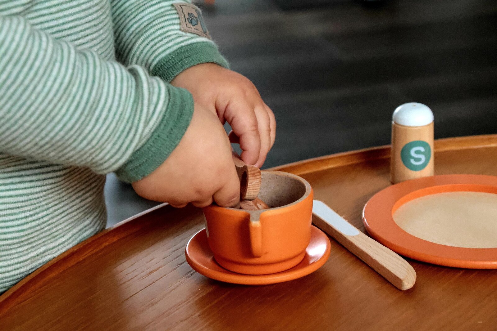Ein kleines Kind in einem gr&uuml;n-wei&szlig; gestreiften Langarmshirt spielt an einem Holztisch mit einem Holzspielzeug-Teeservice. Es h&auml;lt einen kleinen Holzl&ouml;ffel und r&uuml;hrt in einer orangefarbenen Tasse mit Unterteller. Auf dem Tisch stehen au&szlig;erdem ein orangefarbener Spielzeugteller, ein Spielzeugmesser aus Holz und ein Streuer mit einem gr&uuml;nen &bdquo;S&ldquo; f&uuml;r Salz. 