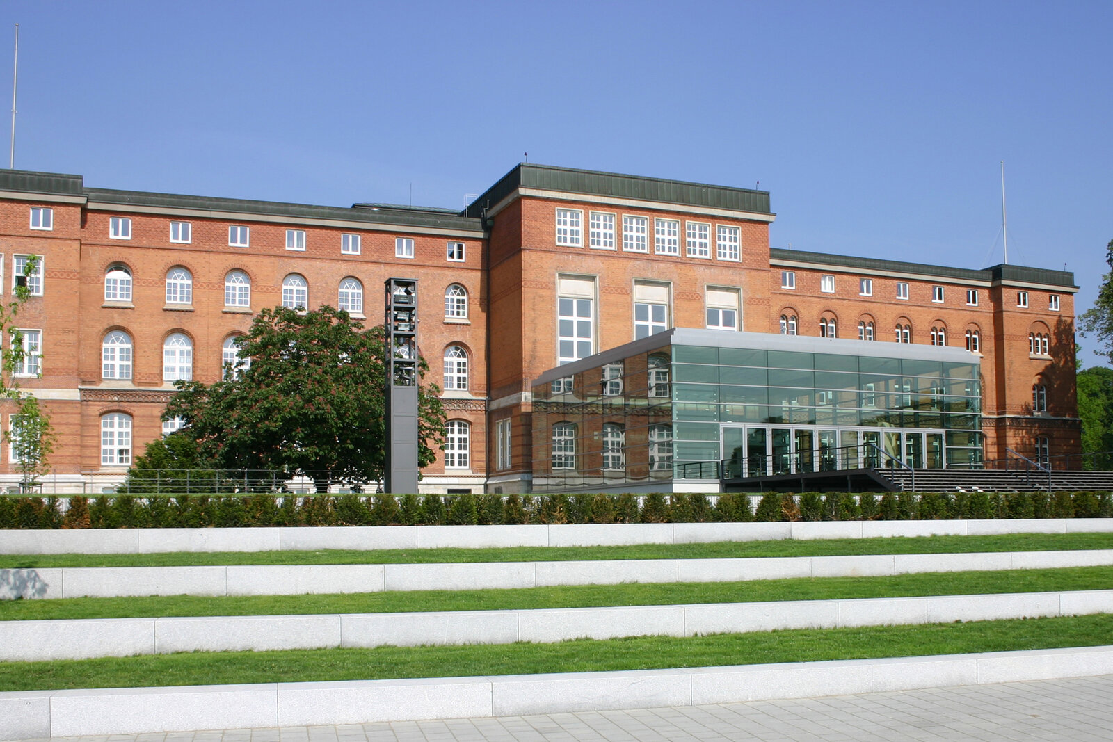 Das Bild zeigt den Landtag von Scleswig-Holstein. Es ist ein sehr gro&szlig;es Geb&auml;ude aus rotem Backstein und es hat einen gro&szlig;en Vorderraum aus Gla&szlig;.