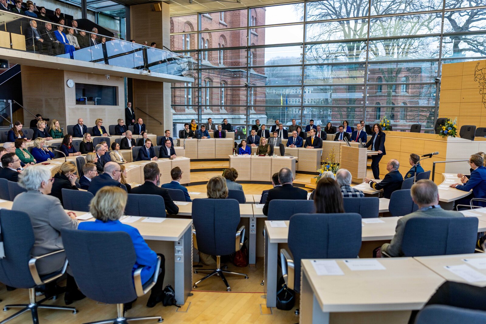 Plenarsaal eines modernen Parlamentsgebäudes mit vielen anwesenden Personen. Die Abgeordneten und Gäste sitzen in einem halbkreisförmig angeordneten Saal mit großen Glasfenstern, durch die ein historisches Backsteingebäude und Bäume zu sehen sind. Eine Rednerin steht am Podium vorne rechts, während die anderen Zuhörenden aufmerksam lauschen.