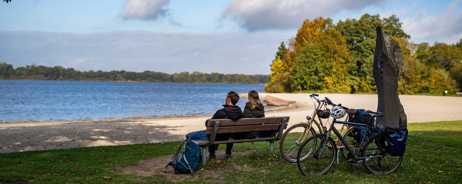Ein Pärchen sitzt auf einer Parkbank an einem See und genießt die Aussicht. Neben ihnen stehen zwei Fahrräder mit Gepäcktaschen und Helmen, die auf eine Radtour schließen lassen. Die Szenerie ist von herbstlich gefärbten Bäumen umgeben und der Himmel zeigt eine Mischung aus blauen und bewölkten Abschnitten. Im Hintergrund erstreckt sich der See mit sandigem Ufer, im Vordergrund liegt ein Rucksack im Gras. Die Atmosphäre ist ruhig und entspannend, perfekt für eine Pause in der Natur.