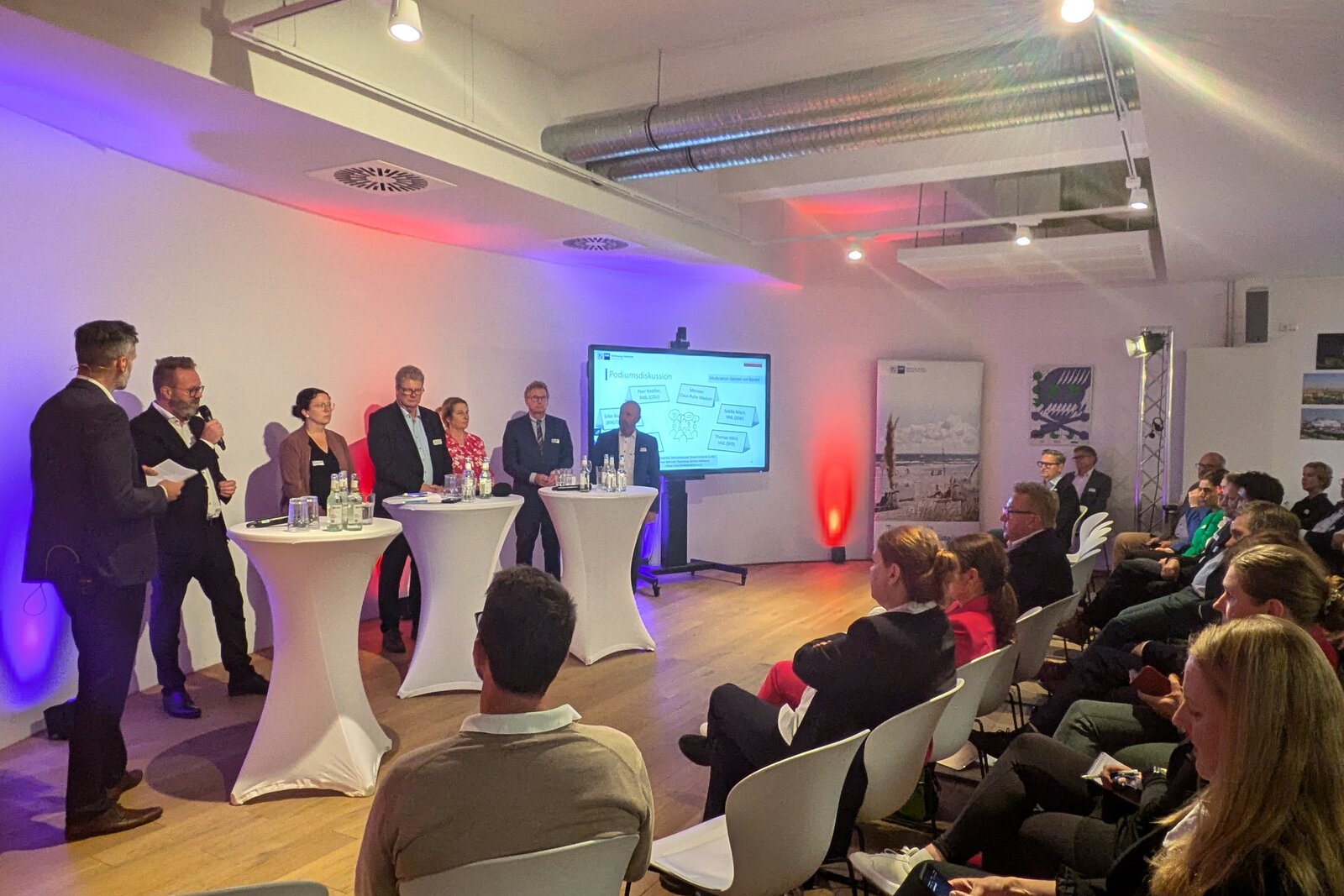 Podiumsdiskussion bei einer Fachveranstaltung: Mehrere Personen stehen an Stehtischen auf einer Bühne, während ein Moderator mit Mikrofon spricht. Eine Präsentationsfolie mit dem Titel 'Podiumsdiskussion' ist im Hintergrund auf einem Bildschirm zu sehen. Das Publikum sitzt aufmerksam in weißen Stühlen, während die Veranstaltung in einem modern gestalteten Raum stattfindet.