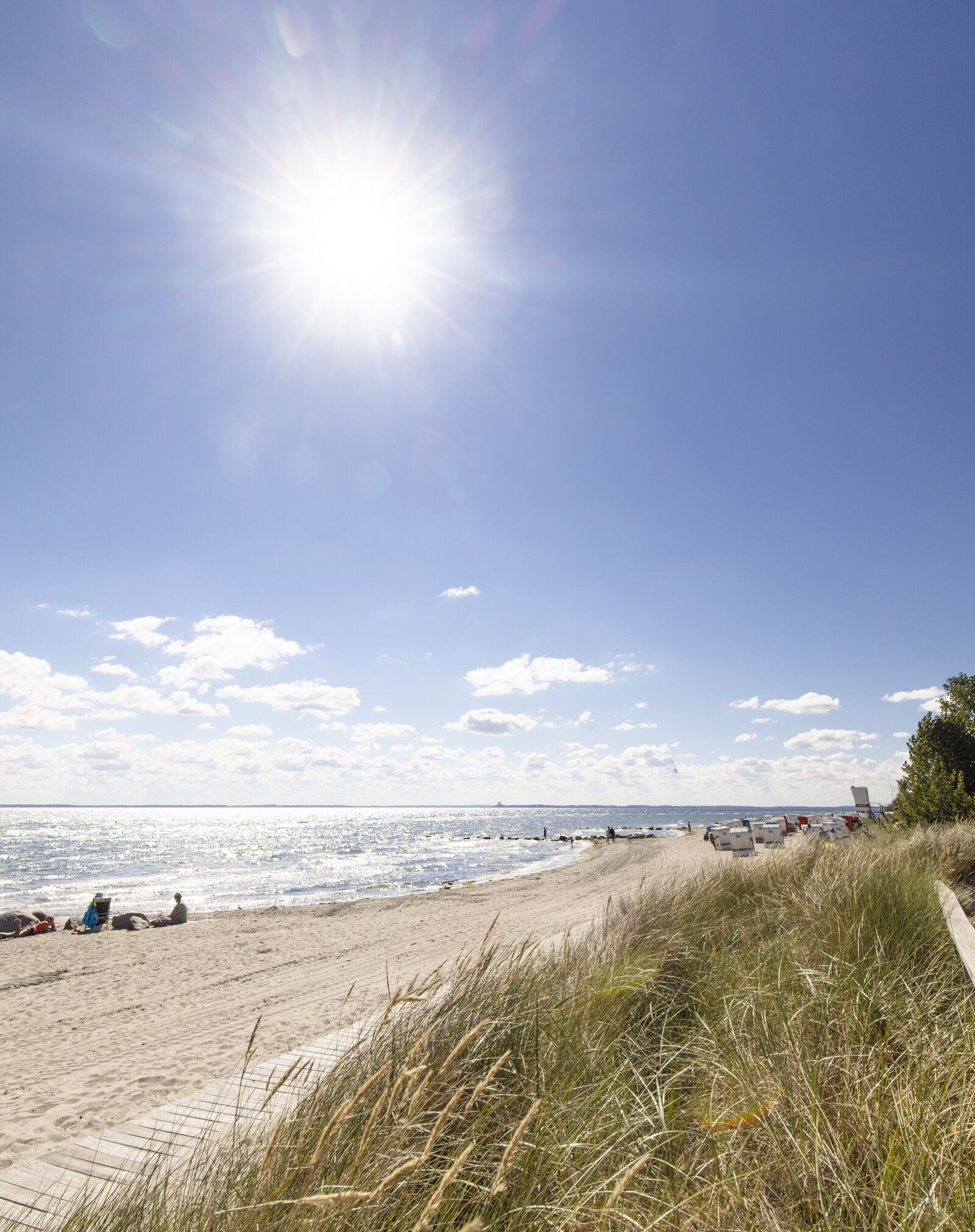 Sonniger Tag am Strand von Rettin an der Ostsee: Im Vordergrund D&uuml;nen mit hohem Gras, im Hintergrund wei&szlig;er Sandstrand mit einigen Menschen, ruhiger Wellengang und mehrere Strandk&ouml;rbe entlang des Ufers. Am Himmel strahlt die Sonne hell &uuml;ber dem klarblauen Himmel mit wenigen wei&szlig;en Wolken.
