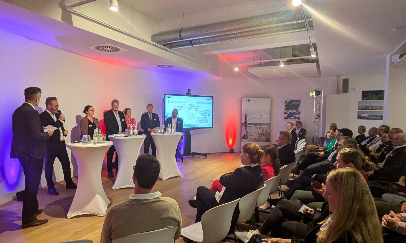 Das Bild zeigt eine lebhafte Veranstaltung in einem modernen, lichtdurchfluteten Raum mit großen Fenstern. Viele Menschen stehen in Gruppen an Stehtischen und unterhalten sich. Einige halten Getränke in der Hand. Die Atmosphäre wirkt entspannt und professionell.