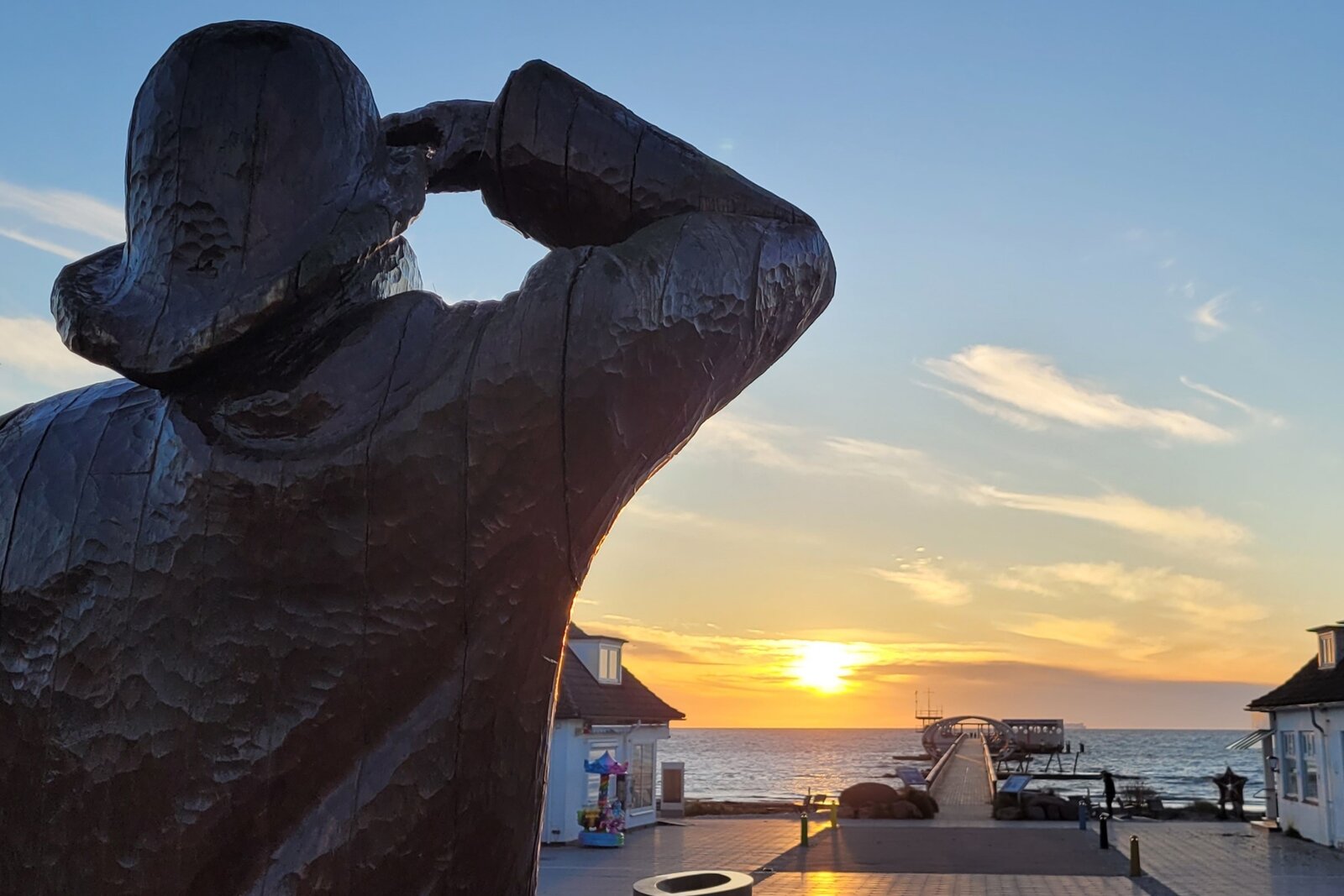 Bild einer K&uuml;stenszene bei Sonnenuntergang: Im Vordergrund eine Holzskulptur eines Fischers mit Hut, der in die Ferne blickt. Dahinter f&uuml;hrt eine Strandpromenade zum Meer, flankiert von Geb&auml;uden auf beiden Seiten. Der Himmel leuchtet in warmen Farben, die Sonne steht knapp &uuml;ber dem Horizont.
