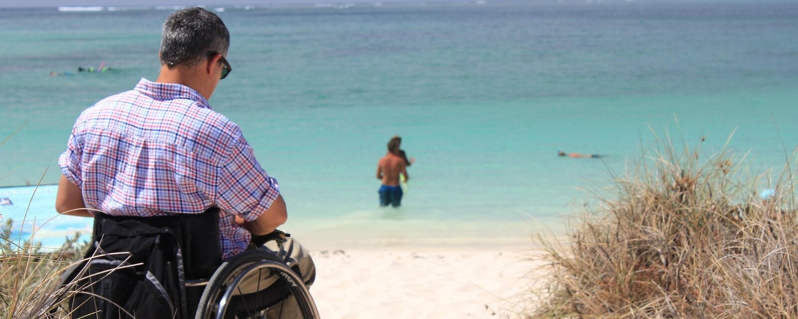 Das Bild zeigt einen Mann im Rollstuhl, der auf einem barrierefreien Holzsteg Richtung Meer schaut. Der Sandstrand f&uuml;hrt sanft in das t&uuml;rkisfarbene Wasser &uuml;ber. Im Hintergrund sind weitere Menschen beim Schwimmen oder Baden. Das Bild vermittelt Barrierefreiheit, Inklusion und Sommerfreude am Strand.