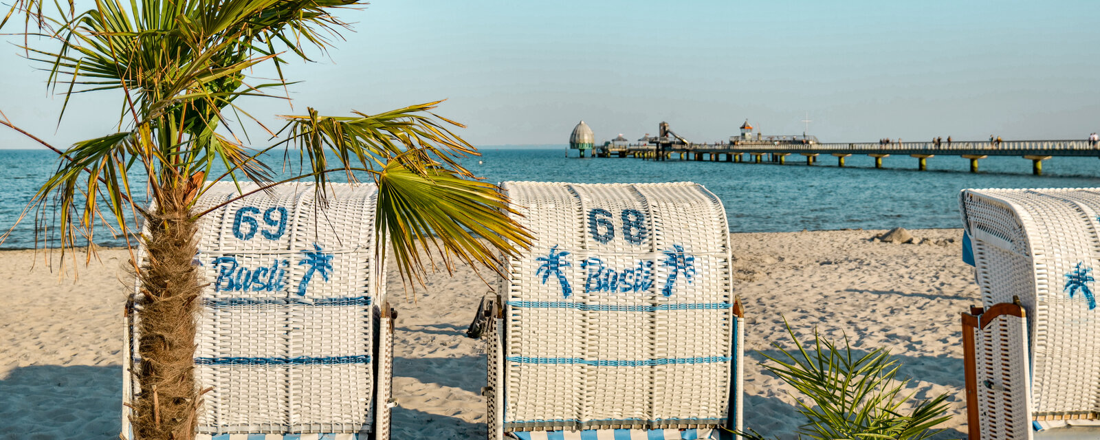 Sommerlicher Strand in Gr&ouml;mitz mit wei&szlig;en Strandk&ouml;rben, einer Palme im Vordergrund und Blick auf die Seebr&uuml;cke Gr&ouml;mitz mit Tauchglocke bei klarem Himmel.
