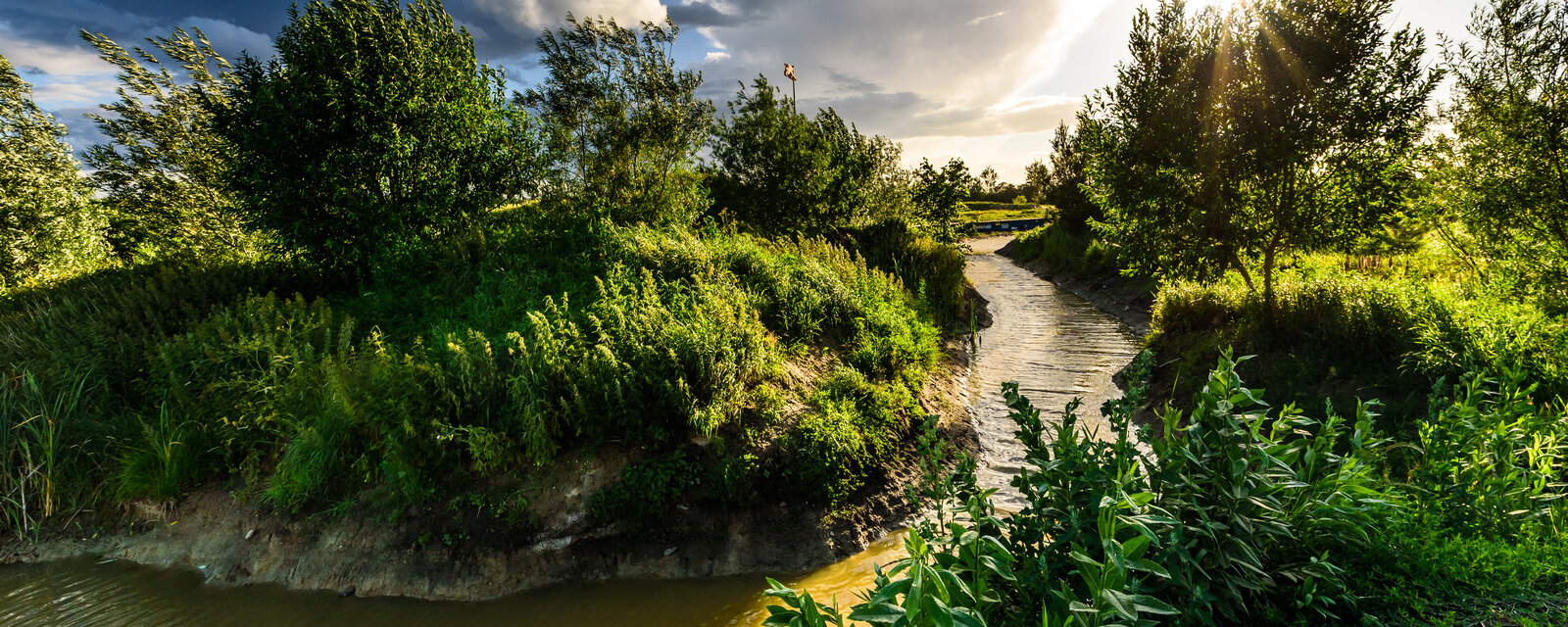 Das Bild zeigt eine naturnahe Landschaft an einem sonnigen Tag. Im Vordergrund flie&szlig;t ein schmaler, gewundener Bach durch &uuml;ppiges gr&uuml;nes Gras und dichte Vegetation. Auf beiden Seiten des Bachs wachsen viele Str&auml;ucher und mittelgro&szlig;e B&auml;ume mit leuchtend gr&uuml;nen Bl&auml;ttern. Der Himmel ist teils blau, teils bew&ouml;lkt, und die Sonne steht tief am Himmel, nahe dem Horizont. Sonnenstrahlen brechen durch die Baumkronen auf der rechten Seite und werfen ein warmes Licht auf die Szene. Im Hintergrund ist auf einem Mast eine wehende Fahne zu sehen.