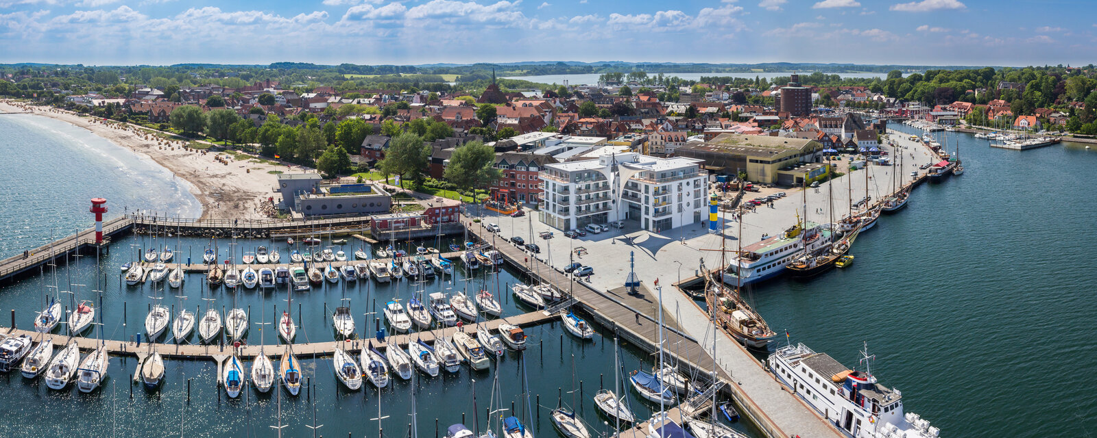Luftaufnahme des Eckernförder Hafens mit zahlreichen Segelbooten, der Strandpromenade, dem feinsandigen Ostseestrand links und der historischen Altstadt im Hintergrund. Im Bild sind auch die Hafenmole mit Leuchtturm, moderne und traditionelle Gebäude sowie grüne Wälder im Umland zu sehen.