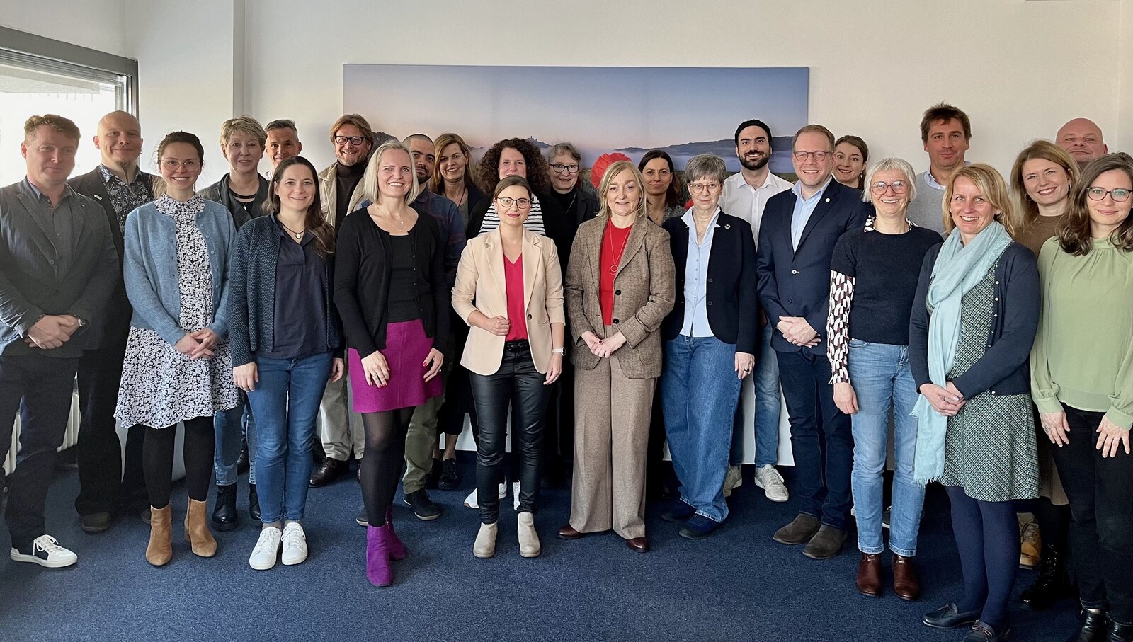 Gruppenfoto einer vielfältigen Arbeitsgruppe in einem modernen Büro mit hellem Licht. Die Personen stehen eng zusammen und lächeln in die Kamera. Sie tragen eine Mischung aus formeller und legerer Kleidung, mit verschiedenen Farben und Stilen. Im Hintergrund ist ein großes Bild mit einer Landschaft zu sehen, das eine entspannte Atmosphäre vermittelt.