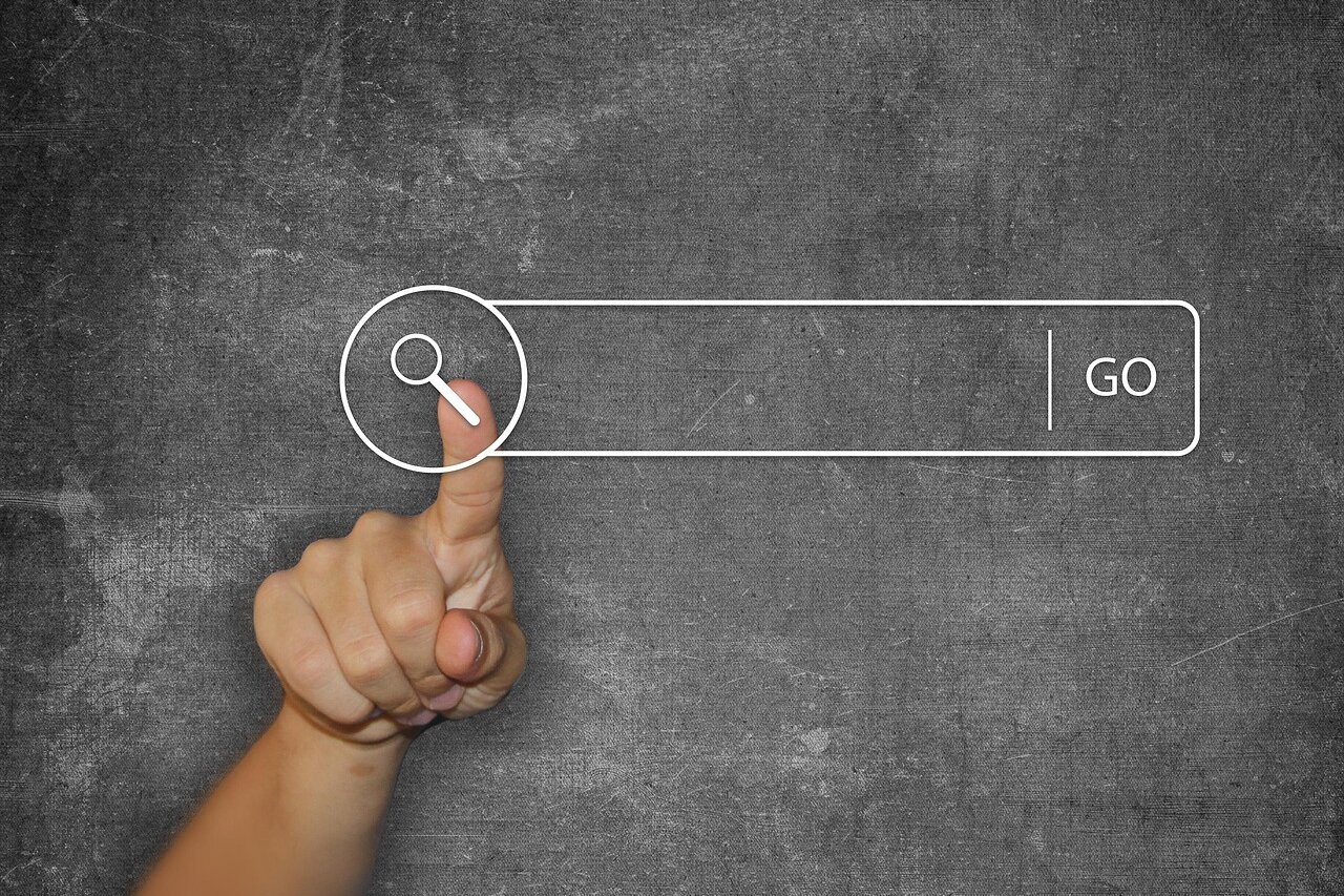 Eine menschliche Hand zeigt mit dem Zeigefinger auf ein digitales Suchfeld, das ein Lupe-Symbol links und die Beschriftung ‚GO‘ rechts enthält, dargestellt auf einem strukturierten grauen Hintergrund.