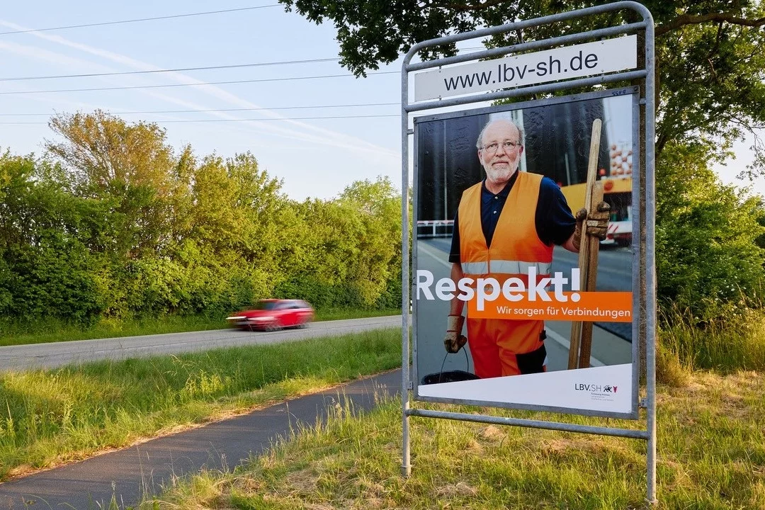 Plakat am Straßenrand mit einem älteren Straßenarbeiter in orangefarbener Warnkleidung und Schaufel. Auf dem Plakat steht: ‚Respekt! Wir sorgen für Verbindungen‘ sowie die Website lbv-sh.de. Im Hintergrund fährt ein Auto auf einer Landstraße vorbei.