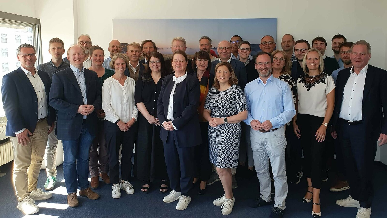 Ein Gruppenfoto von etwa 28 Personen in einem hellen Büro mit großen Fenstern. Die Personen stehen eng beieinander, lächeln und blicken in die Kamera. Sie tragen eine Mischung aus geschäftlicher und legerer Kleidung, oft in Blau-, Schwarz- und Weißtönen. Die Atmosphäre wirkt freundlich und professionell