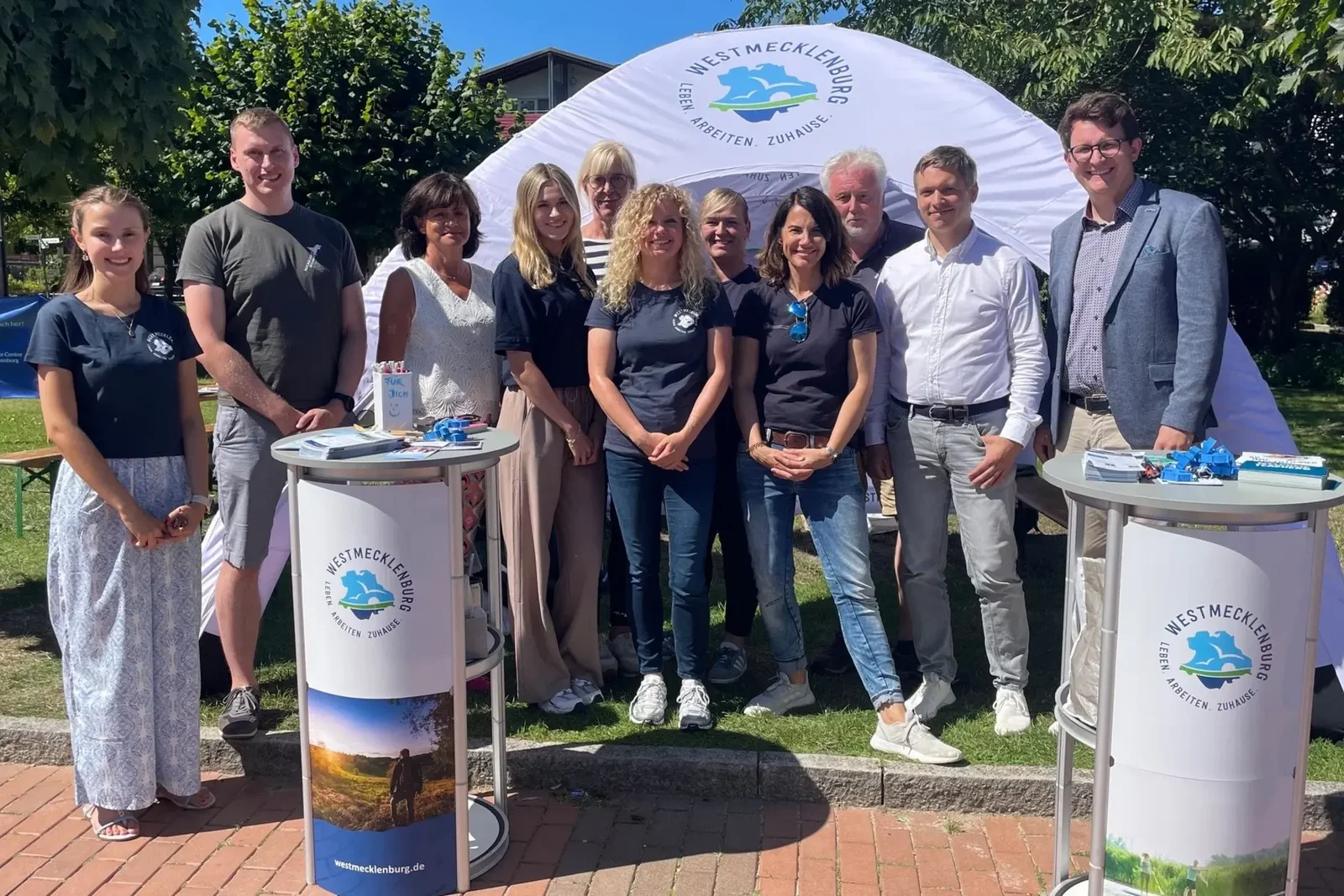 Gruppenfoto des Teams der Urlauberaktionstage 2024 an einem Infostand bei sonnigem Wetter. Im Hintergrund ist ein weißer Pavillon mit dem Logo sichtbar.