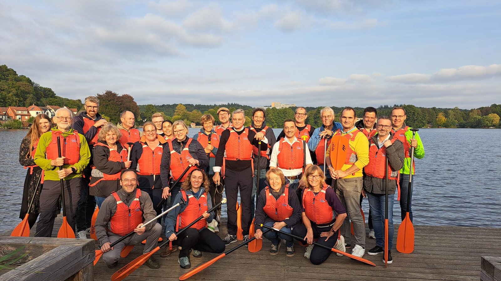 Eine Gruppe von etwa 25 Personen steht auf einem Holzsteg an einem See, umgeben von grüner Natur und kleinen Häusern im Hintergrund. Sie tragen rote Schwimmwesten und halten Paddel in der Hand, bereit für eine gemeinsame Paddeltour. Die Gruppe wirkt fröhlich und motiviert, das Wetter ist sonnig mit einigen Wolken am Himmel. Die Stimmung ist entspannt und gemeinschaftlich.