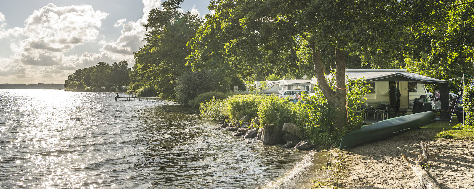 Sonniger Nachmittag am Seeufer eines Campingplatzes. Rechts im Bild stehen unter großen, schattenspendenden Bäumen mehrere Wohnmobile und Zelte. Vor einem Wohnwagen liegt ein grünes Kanu auf dem Boden. Im Vordergrund verläuft ein schmaler Sandstrand entlang des Wassers, das sanft an das Ufer schwappt. Links spiegelt die Sonne auf der Wasseroberfläche. Im Hintergrund führt ein schmaler Steg in den See hinaus, wo eine Person steht.