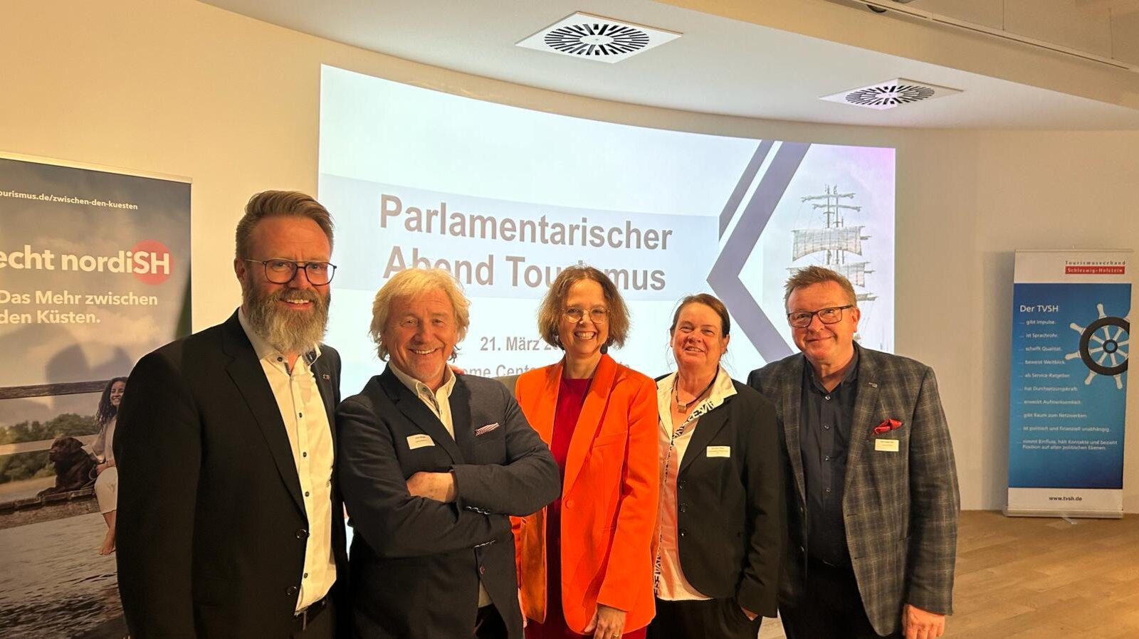 Fünf Personen stehen lächelnd nebeneinander bei einer Veranstaltung mit dem Titel „Parlamentarischer Abend Tourismus“ am 21. März. Im Hintergrund sind zwei Roll-Ups mit Logos und Slogans des Tourismusverbandes Schleswig-Holstein sowie der nordiSH-Kampagne zu sehen. Die Personen tragen überwiegend Businesskleidung und tragen Namensschilder.