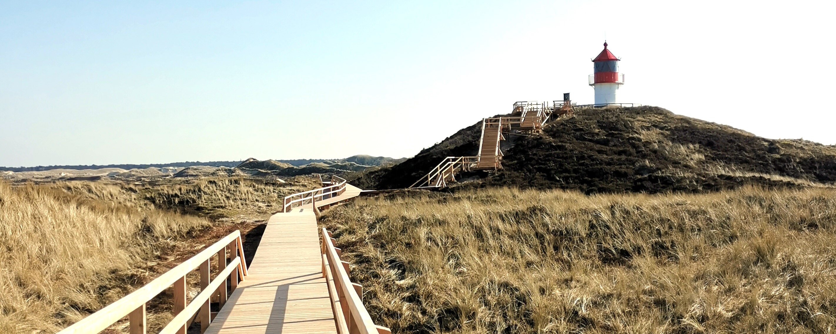 Auf dem Foto ist im Vordergrund ein Bohlenweg zu sehen, der zum Leuchtturm Quermarkenfeuer in Amrum führt.