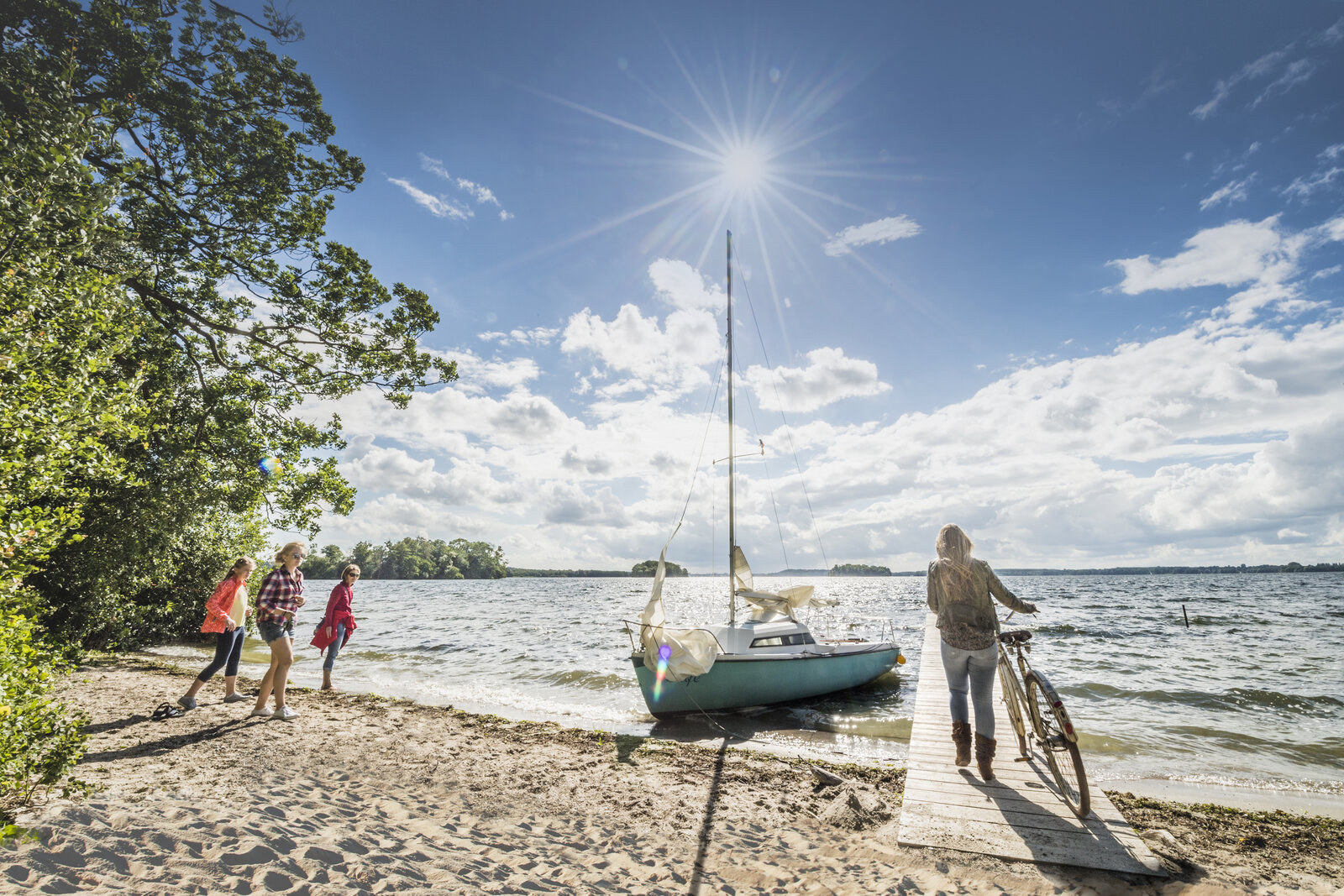 Eine sonnige Sommerlandschaft am Ufer eines Sees: Vier Menschen, darunter Kinder, stehen am Sandstrand neben grünen Bäumen, während eine Frau mit einem Fahrrad auf einem Holzsteg zu einem kleinen Segelboot geht. Der Himmel ist blau mit einigen Wolken, die Sonne scheint strahlend über dem Wasser.