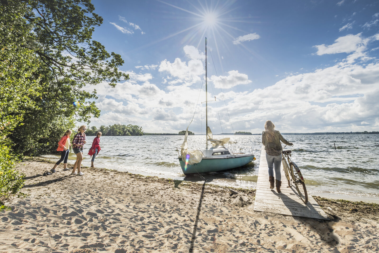 Eine sonnige Sommerlandschaft am Ufer eines Sees: Vier Menschen, darunter Kinder, stehen am Sandstrand neben grünen Bäumen, während eine Frau mit einem Fahrrad auf einem Holzsteg zu einem kleinen Segelboot geht. Der Himmel ist blau mit einigen Wolken, die Sonne scheint strahlend über dem Wasser.