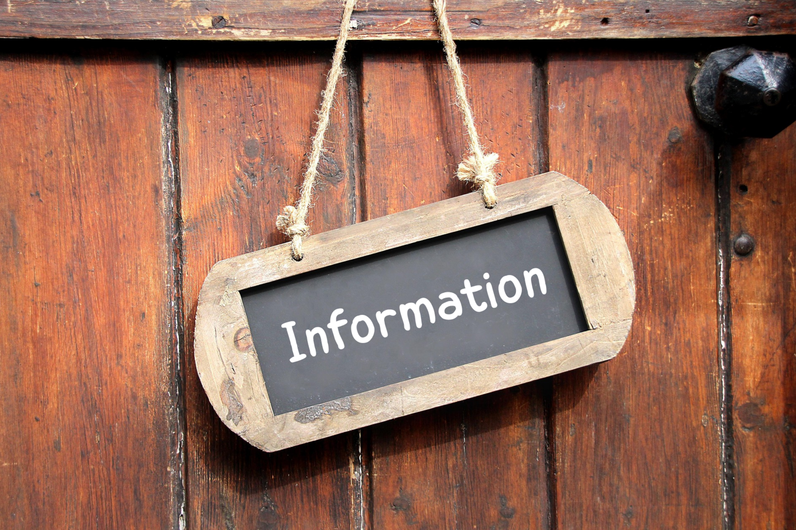 Ein hölzernes Schild mit der Aufschrift ‚Information‘ hängt an zwei Seilen vor einer braunen Holztür.