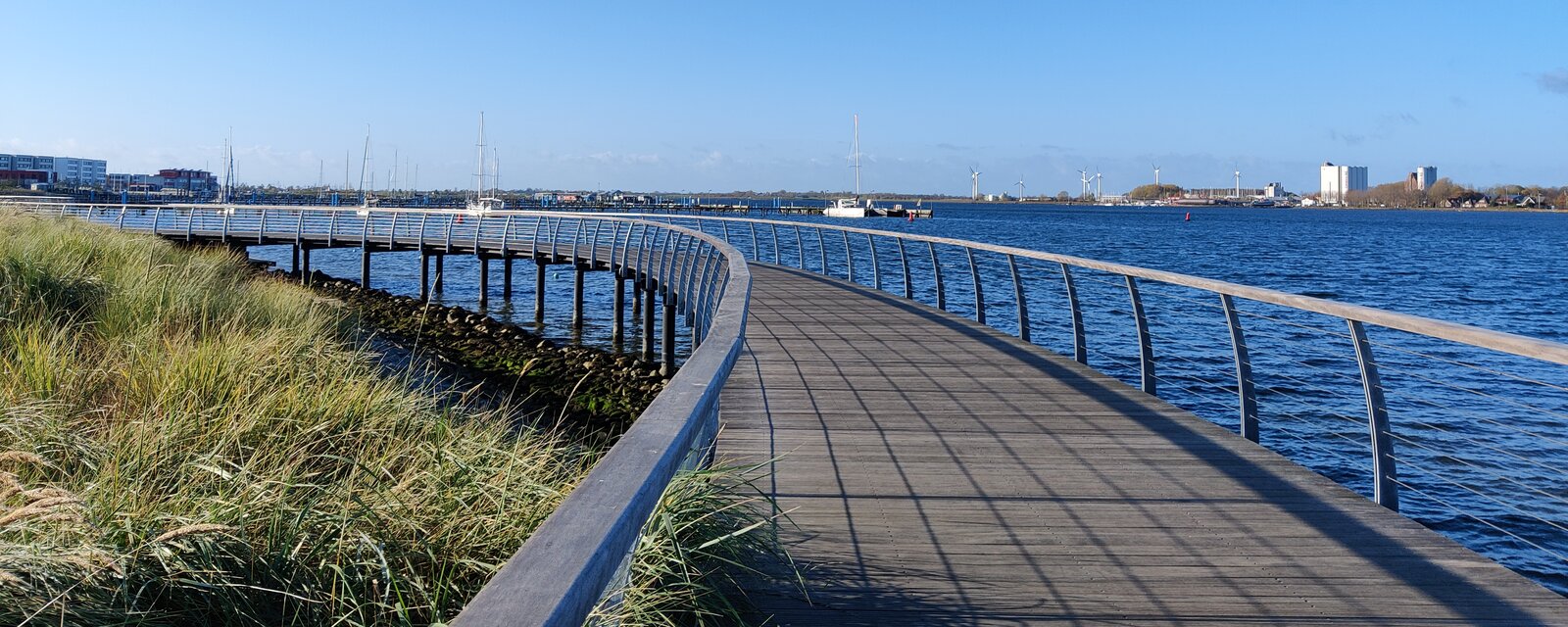 Das Bild zeigt eine moderne, leicht geschwungene Holzpromenade mit Metallgel&auml;nder, die entlang eines Gew&auml;ssers verl&auml;uft und auf Stelzen &uuml;ber das Wasser f&uuml;hrt. Links im Bild w&auml;chst hohes Gras am Ufer, w&auml;hrend rechts das blaue Wasser sanft an die Promenade grenzt. Im Hintergrund sind Segelboote, Geb&auml;ude und Windr&auml;der zu sehen. Der Himmel ist klar und blau, die Szene wirkt ruhig und sonnig &ndash; ideal f&uuml;r einen Spaziergang am Wasser.