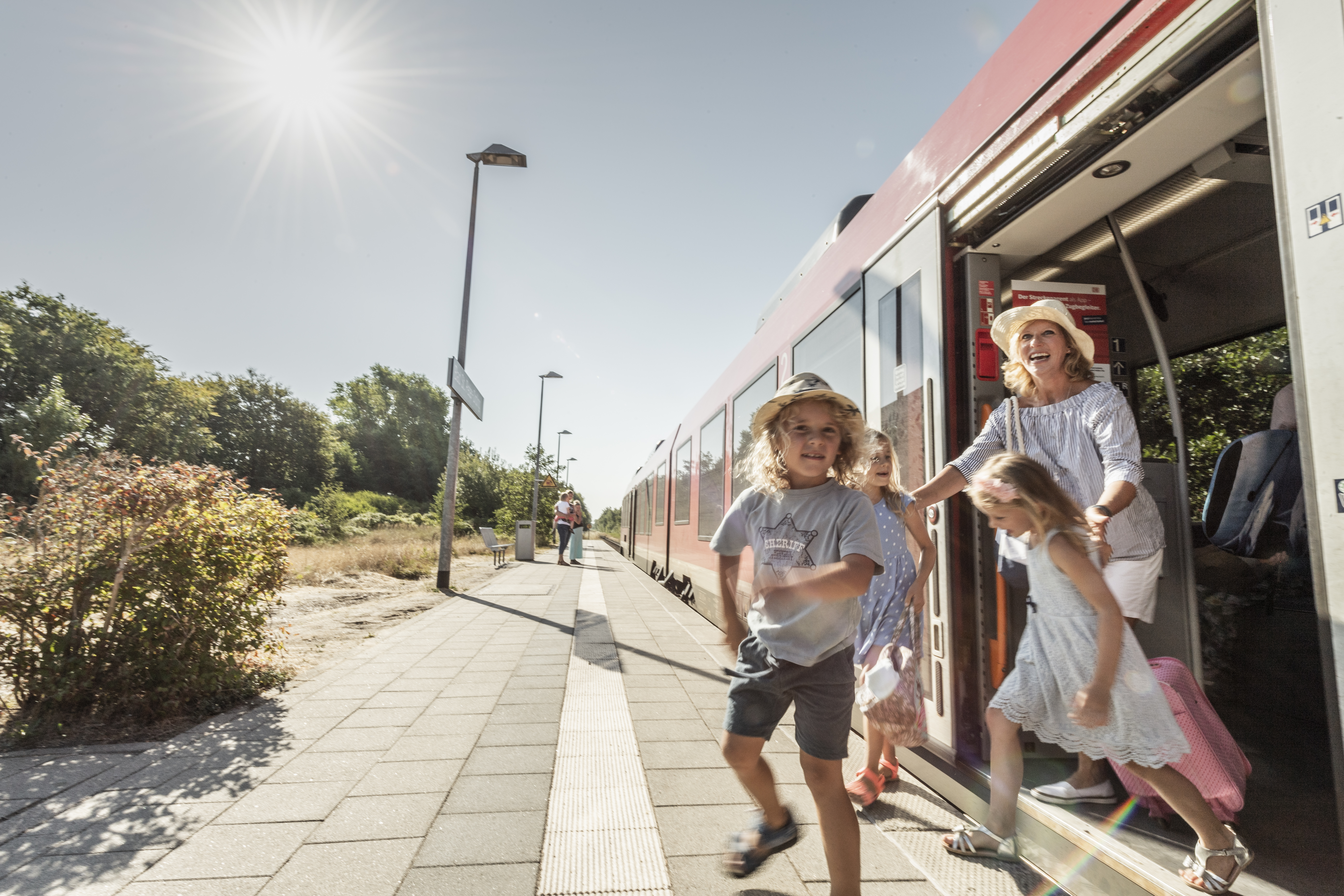 Eine Familie steigt an einem sonnigen Tag auf einem Bahnsteig aus einem roten Zug. Ein Junge läuft fröhlich voraus, die Mutter mit ihren Kindern folgt lächelnd. Der Bahnsteig ist von grüner Vegetation umgeben und die Sonne scheint vom klaren blauen Himmel.