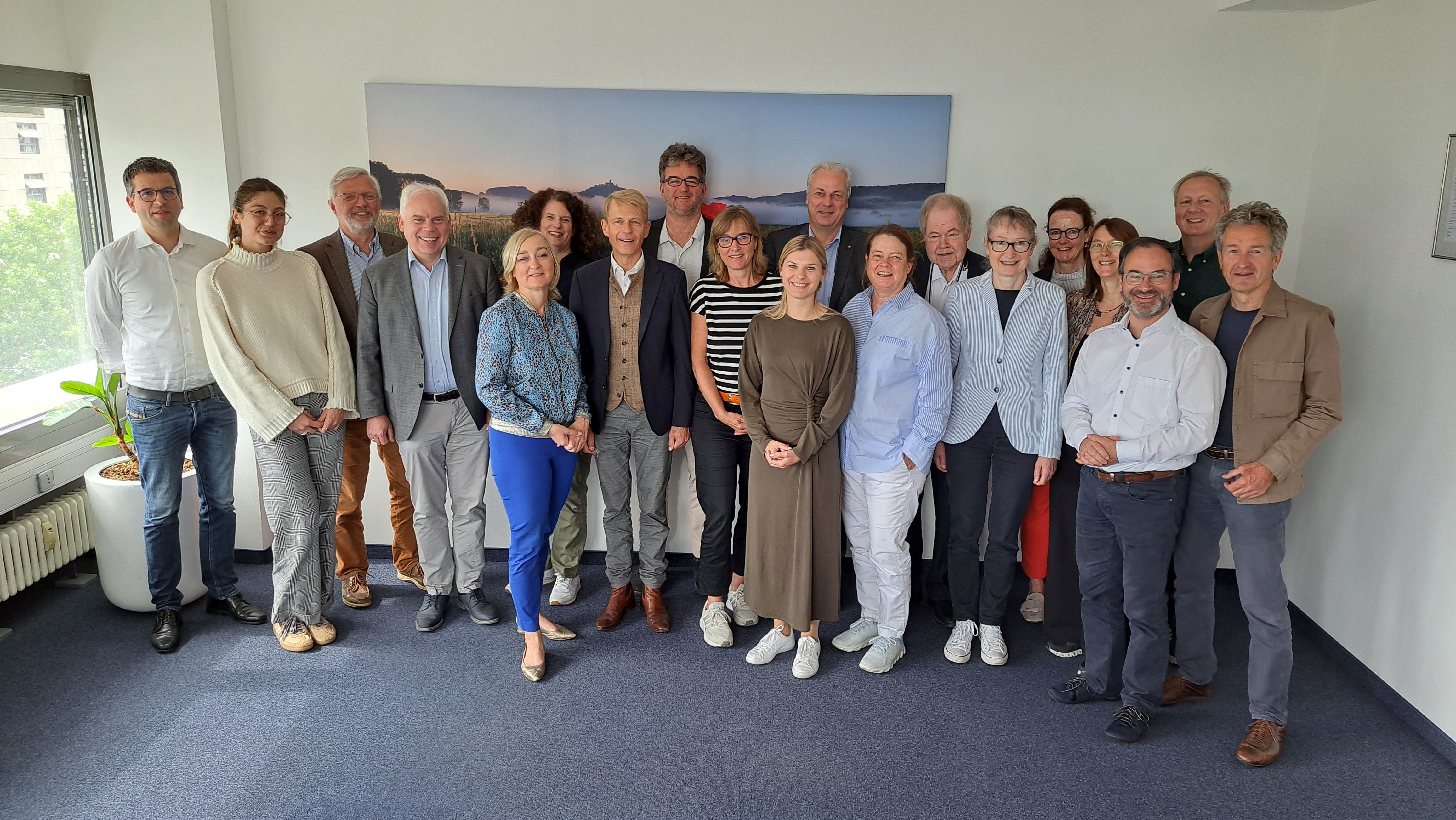 Eine Gruppe von 19 Personen steht in einem Büro vor einer weißen Wand mit einem großen Landschaftsbild. Die Personen sind in einer Mischung aus formeller und legerer Kleidung gekleidet, einige tragen Anzüge, andere Pullover oder Blusen. Viele haben freundliche Gesichtsausdrücke und lächeln in die Kamera. Im Hintergrund ist ein Fenster mit einer Zimmerpflanze zu sehen. Die Atmosphäre wirkt professionell und zugleich entspannt.